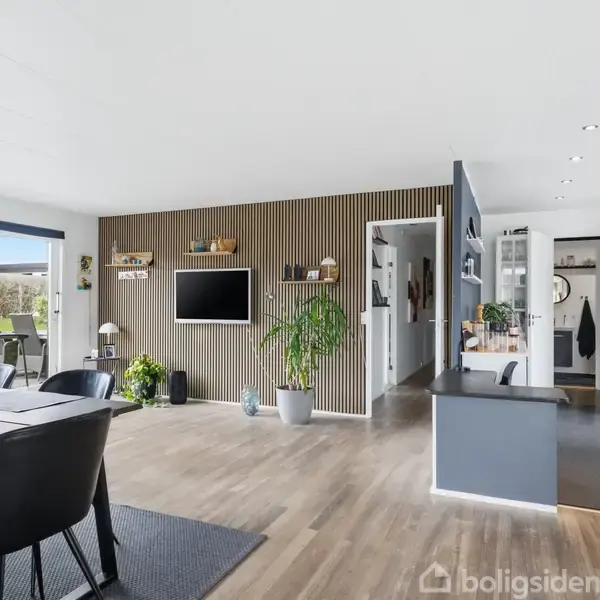 Spisebord med sorte stole står i en moderne stue med trægulv. Et TV hænger på en trævæg. Køkken er synligt til højre, udendørs område til venstre.