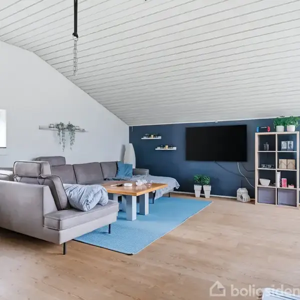 Stue med gråt hjørnesofa står på lyseblåt tæppe. TV hænger på mørk blå væg. Hylde med bøger og planter til højre. Trævindue og loft. Bord i midten.