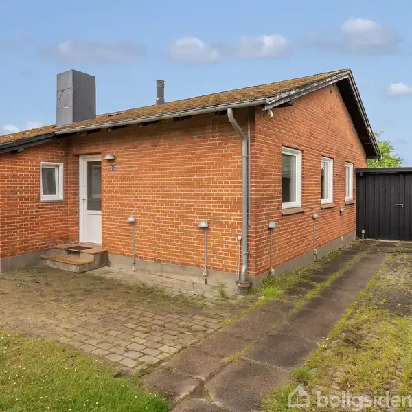 En murstensbygning med rød facade og hvid dør står ved en belagt indkørsel. Græsplæne og buske omgiver huset. Der ses vinduer på siden og en sort skraldespand ved siden af.