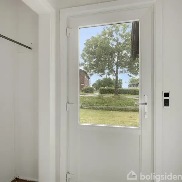 Dør med glasrude åbner ud mod en have med græsplæne og træer. Indendørs ses en hvid væg og en lille garderobe med bøjlestang. Ingen tekst fundet.