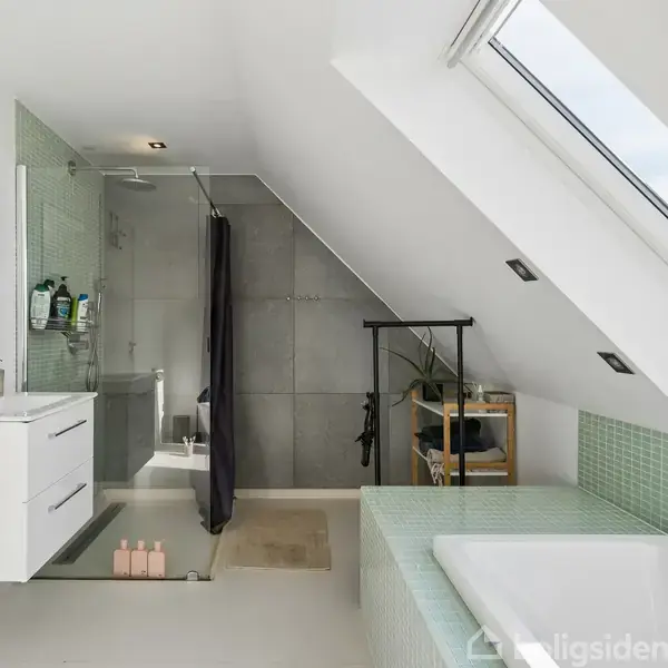 Badeværelse med skråloft; brusekabine med glasvæg, håndvask med spejl, og badekar foran et stort ovenlysvindue; lyse og moderne omgivelser; grønne fliser ved badekar.