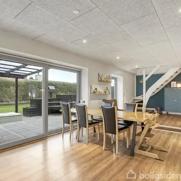 Et spisestuebord med stole står i et moderne rum med trægulv. Store glasdøre fører ud til en terrasse og have. Trappen fører til en øvre etage.