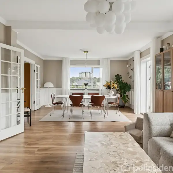 Stue med sofabord og sofa i forgrunden, spisebord med stole i midten, store vinduer, lysestander og grønne planter i baggrunden, trægulv og lyse vægge.