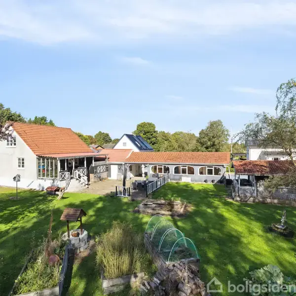 Et hvidt hus med røde tage står i en grøn have med forskellige beplantninger. Solen skinner, og omgivelserne er landlige med klar blå himmel.