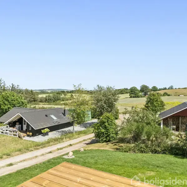 Et sort sommerhus med terrasse står stille i et frodigt, landligt område omgivet af træer og marker. Himlen er klar og blå. En grusvej snor sig forbi huset.