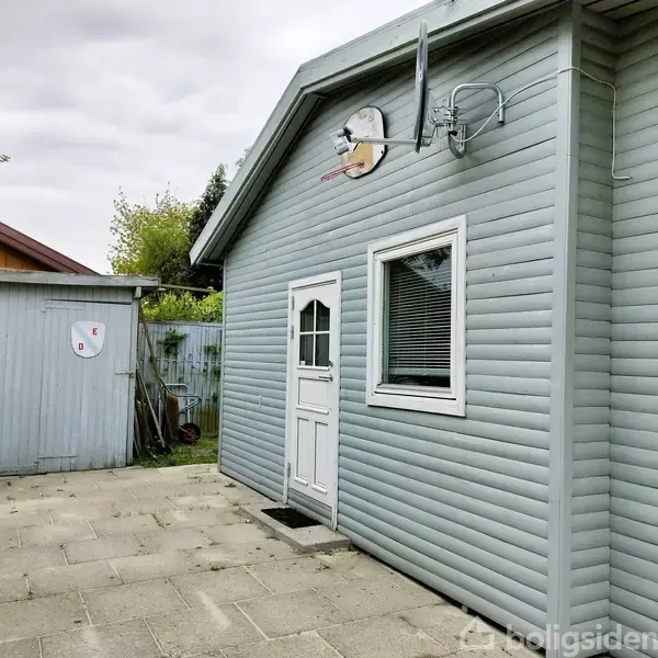 Gråt, træbeklædt hus står stille på en flisebelagt terrasse ved siden af en mindre skurbygning. Satellitantenne monteret på husets væg. Omkranset af grøn vegetation.