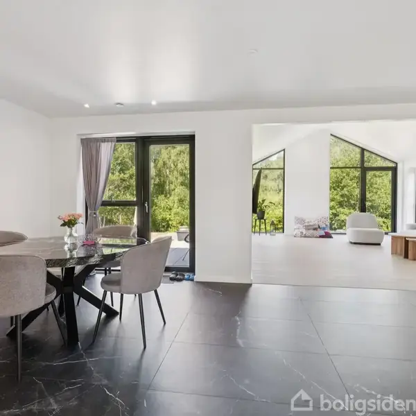 Rundt spisebord med stole står i spisestue med mørkt marmorgulv; fra spisestue ses åben forbindelse til stue med lyse møbler og store vinduer ud mod grønne omgivelser.