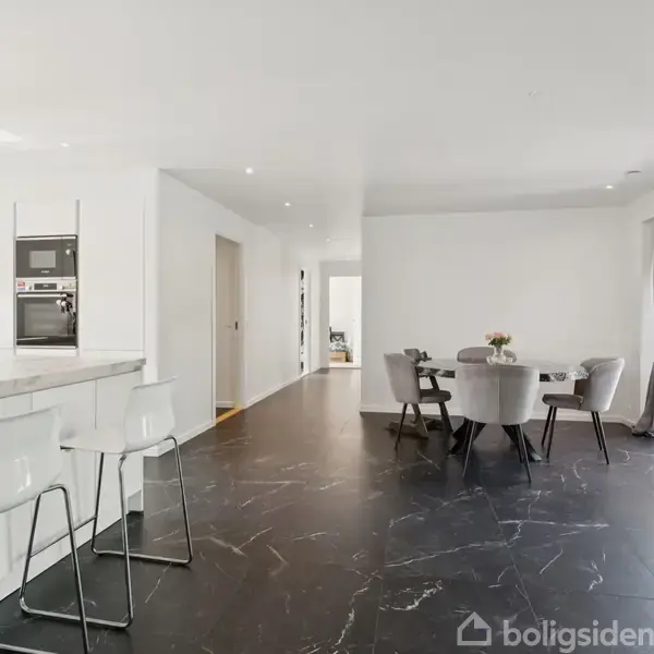 Køkken-spiseområde med hvide vægge og loft, mørke marmorlignende gulve. Køkkenet har en bardisk med hvide stole. Ved siden, et spisebord med grå stole, omgivet af vinduer og glasdør.