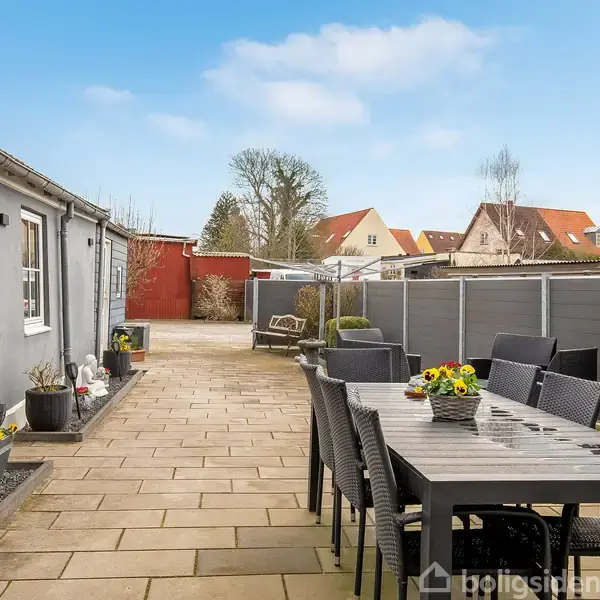 En lang, udendørs terrasse med et sæt sorte havemøbler står stille ved siden af et gråt hus. Omkring er der hegn og andre bygninger i baggrunden under en klar blå himmel.
