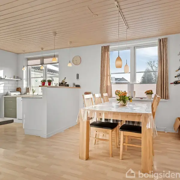 En lys spisestue med et træbord dekoreret med blomster. Seks stole står omkring bordet. Baggrund har store vinduer, tilkoblet køkken og trægulve. En vinholder hænger på væggen.