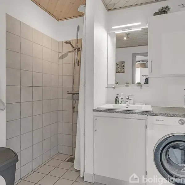 Badeværelse med brusekabine til venstre, vaskemaskine under håndvask og skab til højre. Rummet har fliser på gulvet og lyst træloft. Toilettet er synligt delvist til venstre.