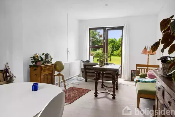 Spisestue med et gammelt bord og stole foran et stort vindue. Rummet er lyst med planter og en sofa i baggrunden. Udenfor ses en grøn have.