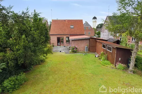 En murstensbygning med rødt tag står i en grøn have med græsplæne og træer. I baggrunden ses et tårn, og til højre er der et træskur.