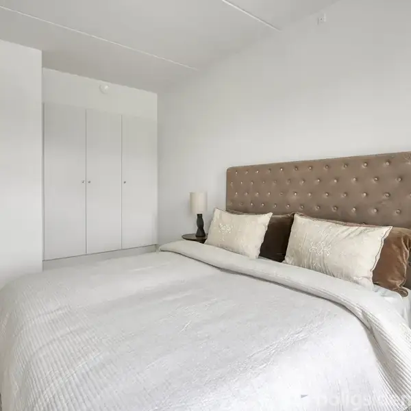 Seng med beige sengegavl er dækket af hvide og brune puder og sengetæppe. To natborde med lamper står på hver side. Værelset har hvidmalede vægge og indbyggede skabe.