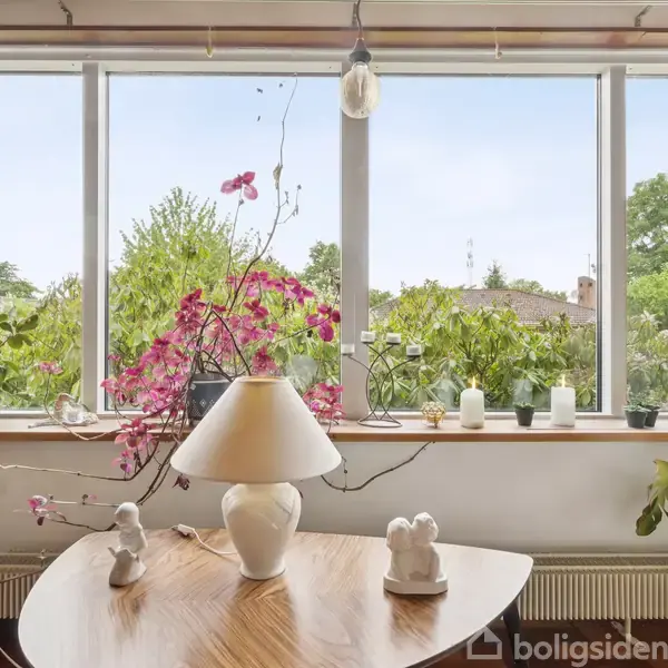 En stue indeholder et bord med en hvid lampe og figurer. Baggrunden viser store vinduer med udsigt til en grøn have. En lyserød blomst står ved vinduet.