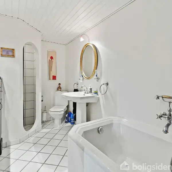 Badeværelse med hvidt flisegulv og vægge, hvor et badekar med armatur står i forgrunden, mens en håndvask med spejl er ved bagvæggen; der hænger malerier på væggen.