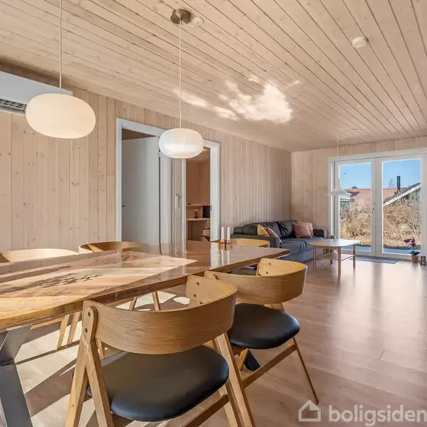 Træspisebord med blomsterdekorationer er omgivet af stole; i baggrunden ses en sofa ved terrassevinduer i et rum med træbeklædte vægge og loft. En airconditionenhed hænger på væggen.