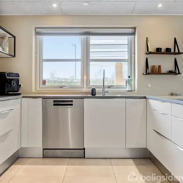 Køkken med hvide skabe og en opvaskemaskine under en stor vask. Kaffemaskine og hylder står på en bordplade. Vindue viser udsigt til naturen udenfor.