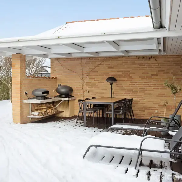 Terrasse med to liggestole og et bord med stole under et halvtag. Sne dækker jorden. En murstensvæg omkranser området, hvor der også er en grill og en lampe.