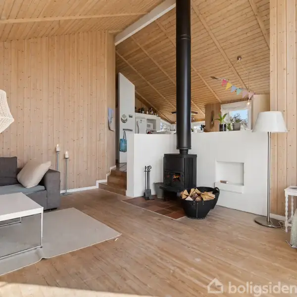Brændeovn i moderne stue opvarmer rummet. Grå sofa, sofabord, lænestol, og gulvlampe skaber hjemlig atmosfære. Lys træbeklædning og åbent køkken i baggrunden. Skilt: "BEACH HOUSE VIBES".