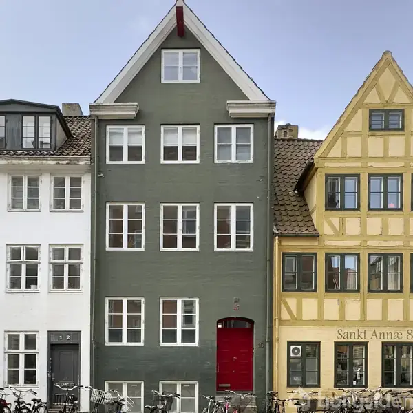 Grå bygning med tre etager, hvidrammede vinduer og en rød dør centreret mellem naboer. Til venstre ses en hvid bygning med nummeret "12," og til højre en gul bindingsværkshus.