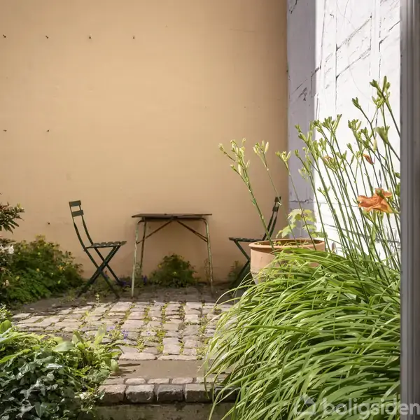 To grønne stole og et lille bord står på en brostensbelagt terrasse omgivet af grønne planter og blomster, foran en lys mur i en lukket gårdhave.