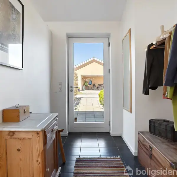 Entré med kommode og knagerække, tøj hænger på bøjler. Dør fører ud til flisebelagt terrasse og carport i baggrunden. Et tæppe ligger på gulvet.