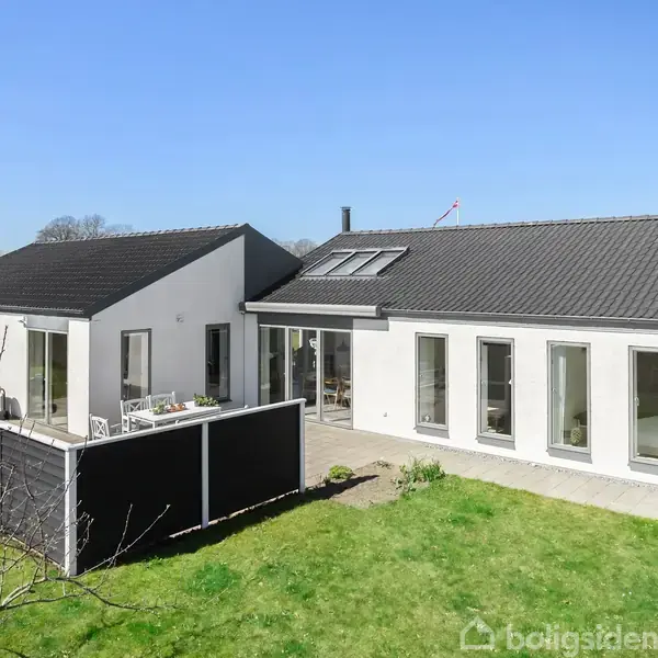 Et hvidt, moderne hus med sort tag står i en have med græsplæne. Store vinduer og en terrasse ses i solskin.