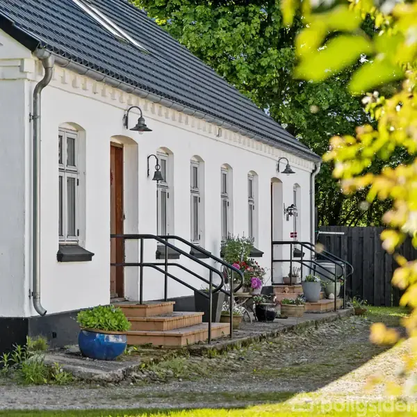Hvidt hus med skråt tag. Trapper fører op til døre; krukker med planter står langs facaden. Grusvej og grøn have omkranser huset, omgivet af frodig vegetation og træer.