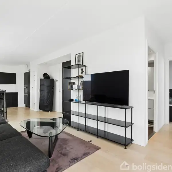 En stue med en sort sofa mod en væg; en fladskærm-tv står på en reol. Et glas-sofabord i forgrunden. Til venstre ses et åbent køkken.