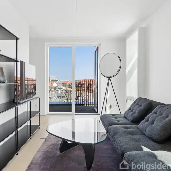 Sofa står ved et glasbord i en lys stue. Store vinduer viser en udendørs balkon og byudsigt. En reol med bøger og pyntegenstande er ved væggen. Ingen tekst.