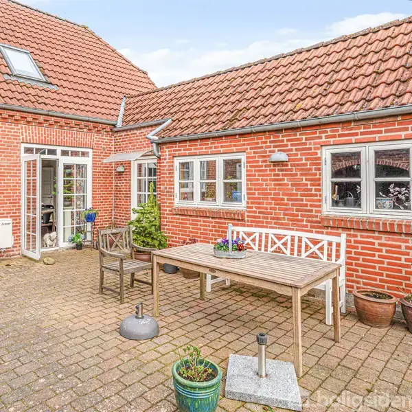 En gårdhave med et træbord og bænkesæt står på en brolagt terrasse foran et rødt murstenshus med hvide vinduer. Planter i krukker omringer området.