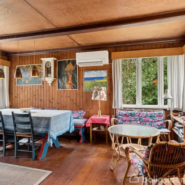 Stue med træbeklædte vægge har spisebord med blå dug og sorte stole til venstre. Til højre ses sofahjørne med rattan møbler. Vinduer viser udsigt til træfyldt område.