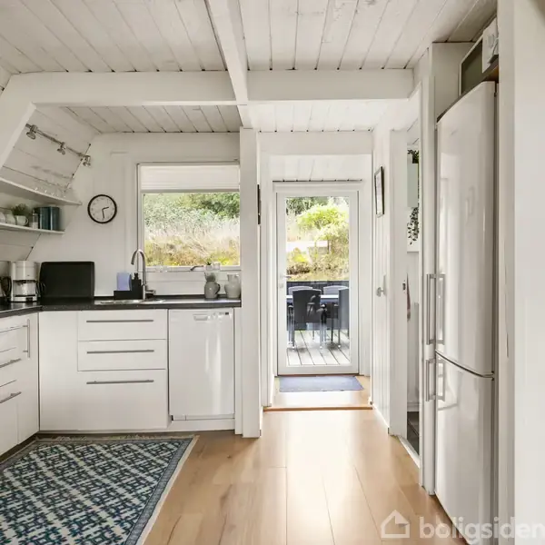 Et hvidt, moderne køkken med indbyggede skabe og apparater. Et tæppe ligger på gulvet, og køkkenet åbner ud mod en terrasse gennem en glasdør, som giver naturligt lys.