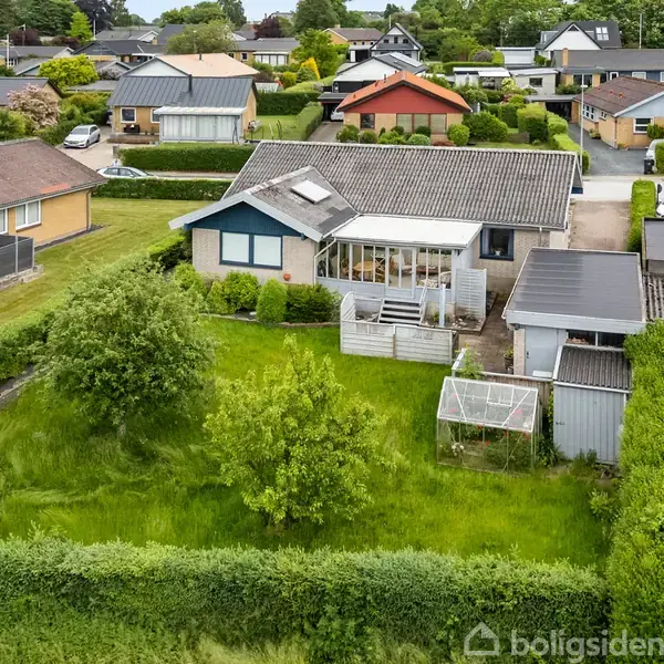 Hus placerer sig i en grøn have med en terrasse og drivhus, omringet af hække og andre huse i et parcelhuskvarter.