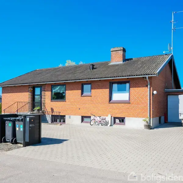 Et murstenshus står stille foran en flisebelagt indkørsel med affaldscontainere til venstre, en cykel lænet mod væggen samt en trailer til højre, omgivet af grøn buskads.
