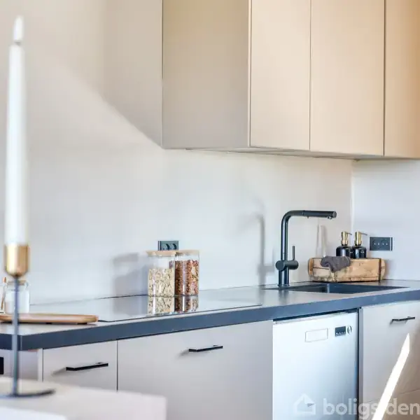 Et moderne køkkenbord med sorte bordplader og hvide skabe. To slanke lysestager står foran. Kryderiglas og køkkenredskaber er placeret ved bordkanten i et velorganiseret rum.