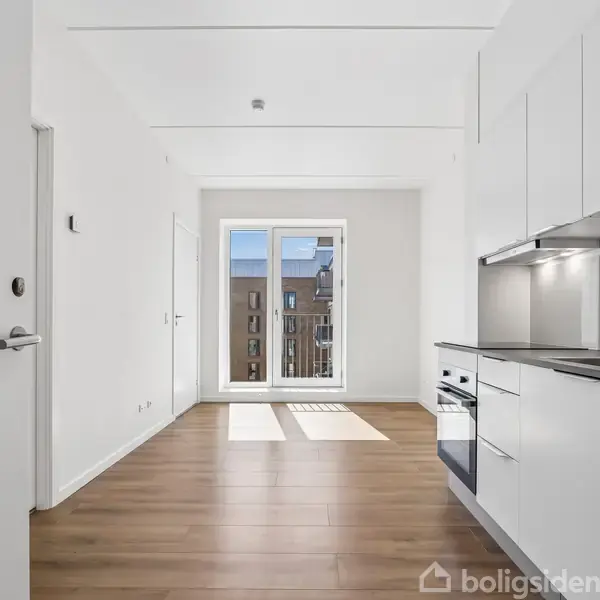 Et lyst køkken med hvide skabe og integrerede hvidevarer; stort vindue med udsigt til en balkon. Trægulv i et moderne, minimalistisk interiør. Ingen tekst fundet.