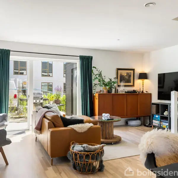 En stue møbleret med en sofa og lænestol centreret omkring et sofabord. Der er en reol med TV til højre, trappe til højre, og glasdøre med grønne gardiner til haven.
