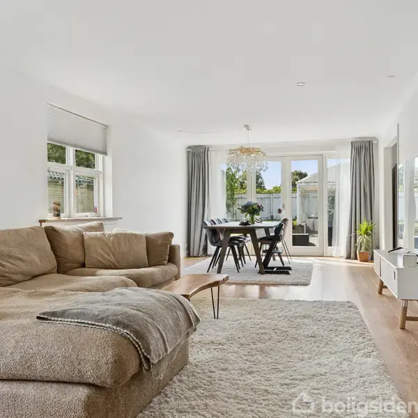 Stor beige sofa står i en moderne stue med trægulv, tæppe og spisebord i baggrunden. Store vinduer og glasdøre giver udsigt til grønne omgivelser.