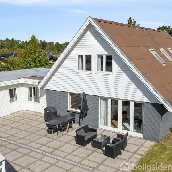 Et hvidt, trekantet taghus med en stor terrasse er omgivet af plæne og træer. Terrassen har sorte udemøbler og en grill, beliggende i en rolig, grøn forstad.