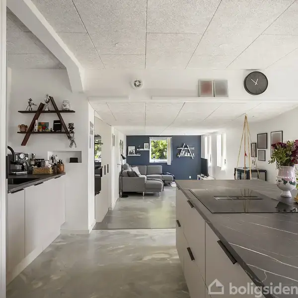 Et moderne køkken, hvor en køkkenø indeholder en vask og knivblok. Rummet har hvide vægge og loft, med et ur på væggen. Stue i baggrunden, blomster på bordet.
