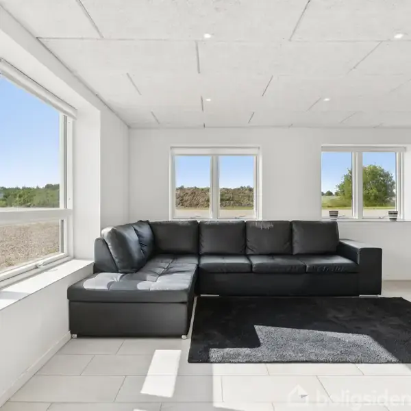 Sort lædersofa står i hjørnet af en lys stue med store vinduer, der slipper masser af dagslys ind. Et sort tæppe ligger foran sofaen på flisegulvet.