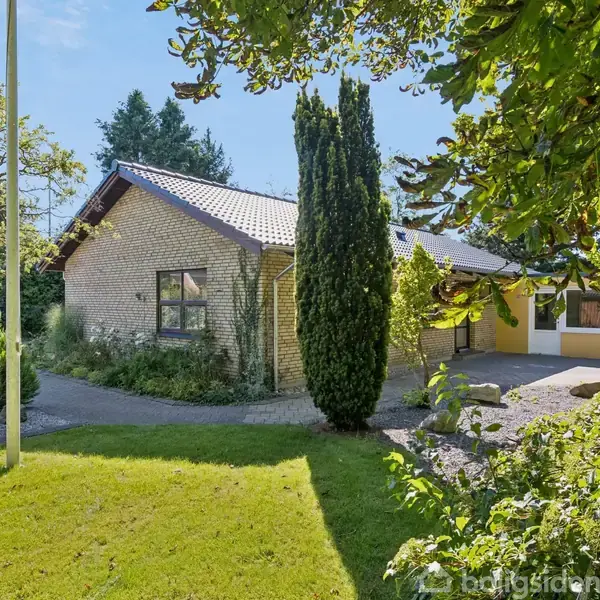 Hus med gult og hvidt murværk omgivet af en velholdt have med træer og buske. Sollys skinner på græsplænen, og der er en asfalteret indkørsel til højre.