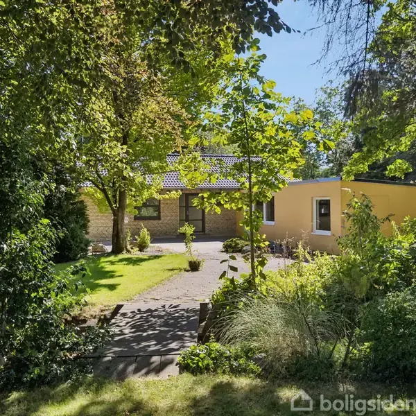 Hus omgivet af en frodig have med træer. Baggrunden viser en lys murstensfacade med en gul tilbygning. Skyggefulde stier fører til huset gennem det grønne område.