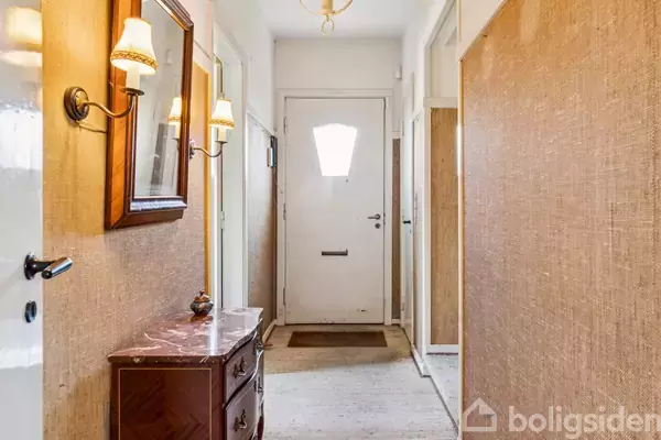 En smal entré med en hvid hoveddør i baggrunden. En kommode med en marmorovertop står til venstre under et spejl og to væglamper. Der er væg-til-væg tæppe på gulvet.