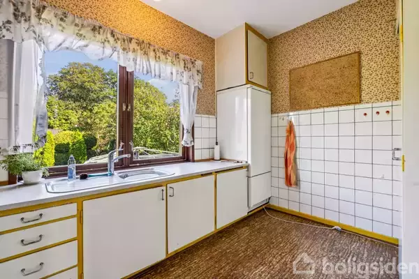 Køkken med hvidt skab, stor vindue med blomstrede gardiner og udsigt til en grøn have. Der er en hvid vask, køleskab og beige vægge med hvidt fliseskjold.