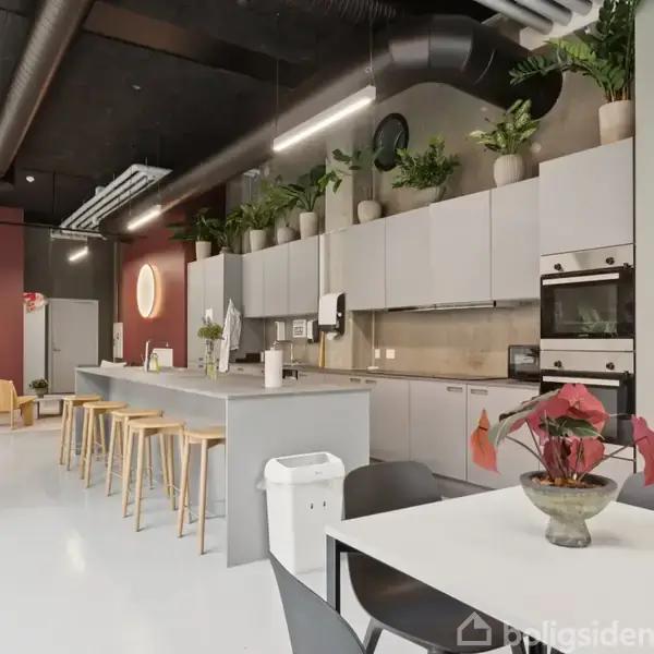 Et moderne køkkenområde med en lang ø med barstole omgivet af skabe og ovne. Området er dekoreret med planter, og en spiseplads med et bord og stole er synlig foran.
