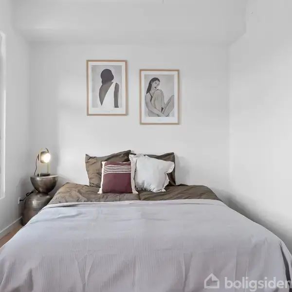 En seng står i et lyst værelse, omgivet af hvide vægge og en lampe på et bord. To indrammede kunstværker hænger over sengen. Ingen tekst er synlig.