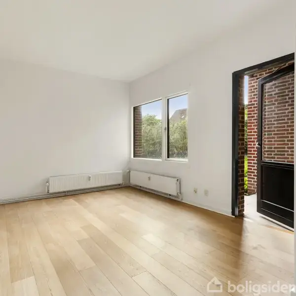 Et tomt rum med trægulv, to vinduer og en åben dør. Væggene er hvide, og der er en radiator under vinduerne. Udenfor ses mursten.